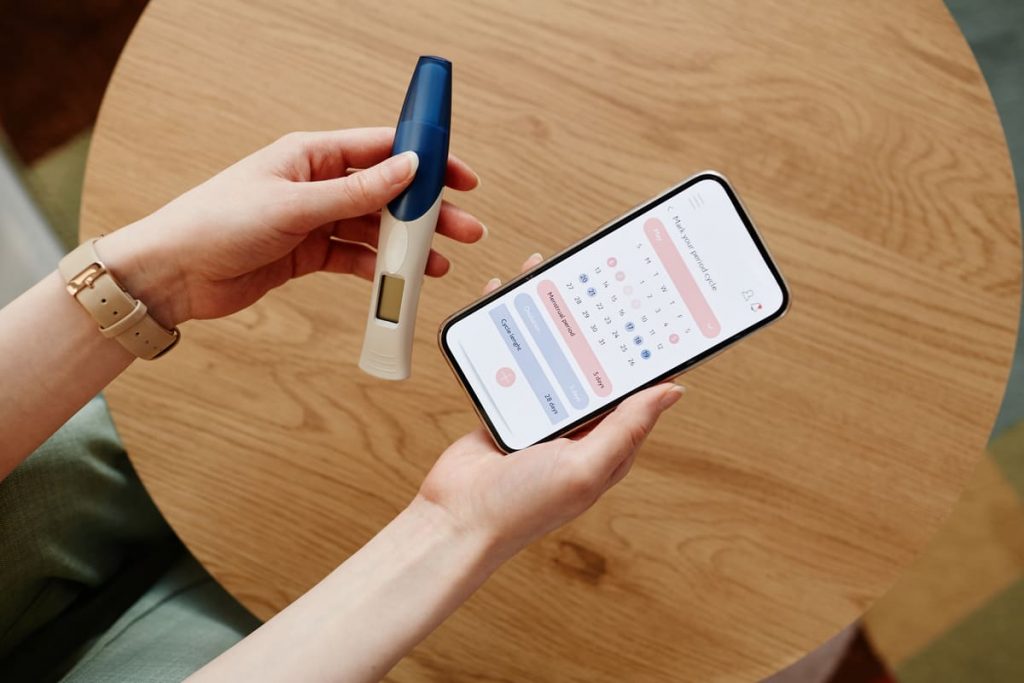 manos sosteniendo un móvil con un calendario menstrual y un test de embarazo preguntándose cómo hacer un bebé